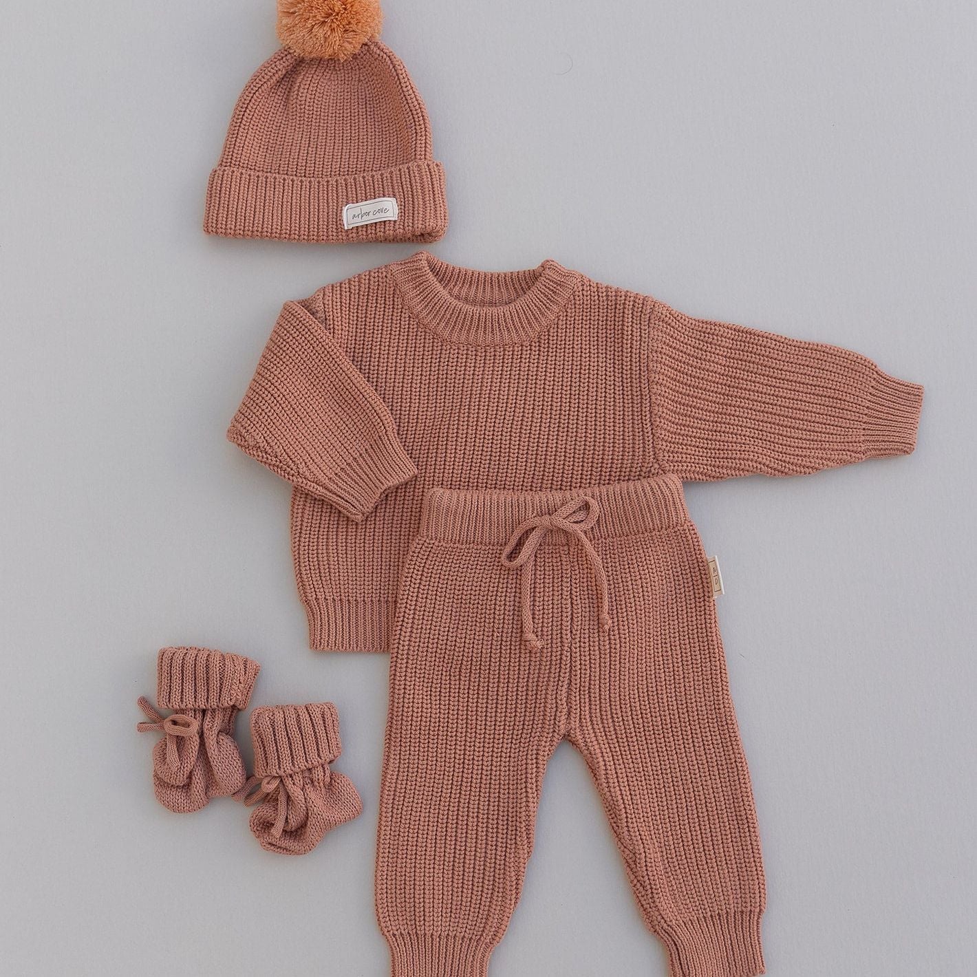 Knitted baby set with sweater, pants, hat, and booties in rust color on a gray background