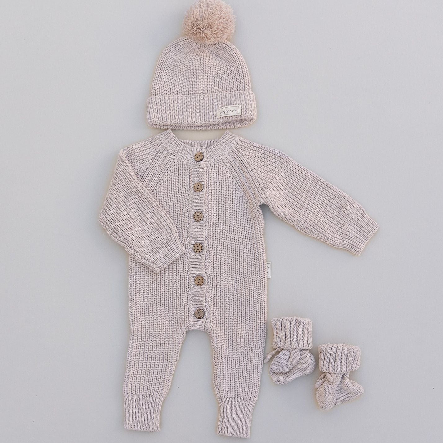 Knitted baby outfit in cream with romper, hat, and socks on a light gray background