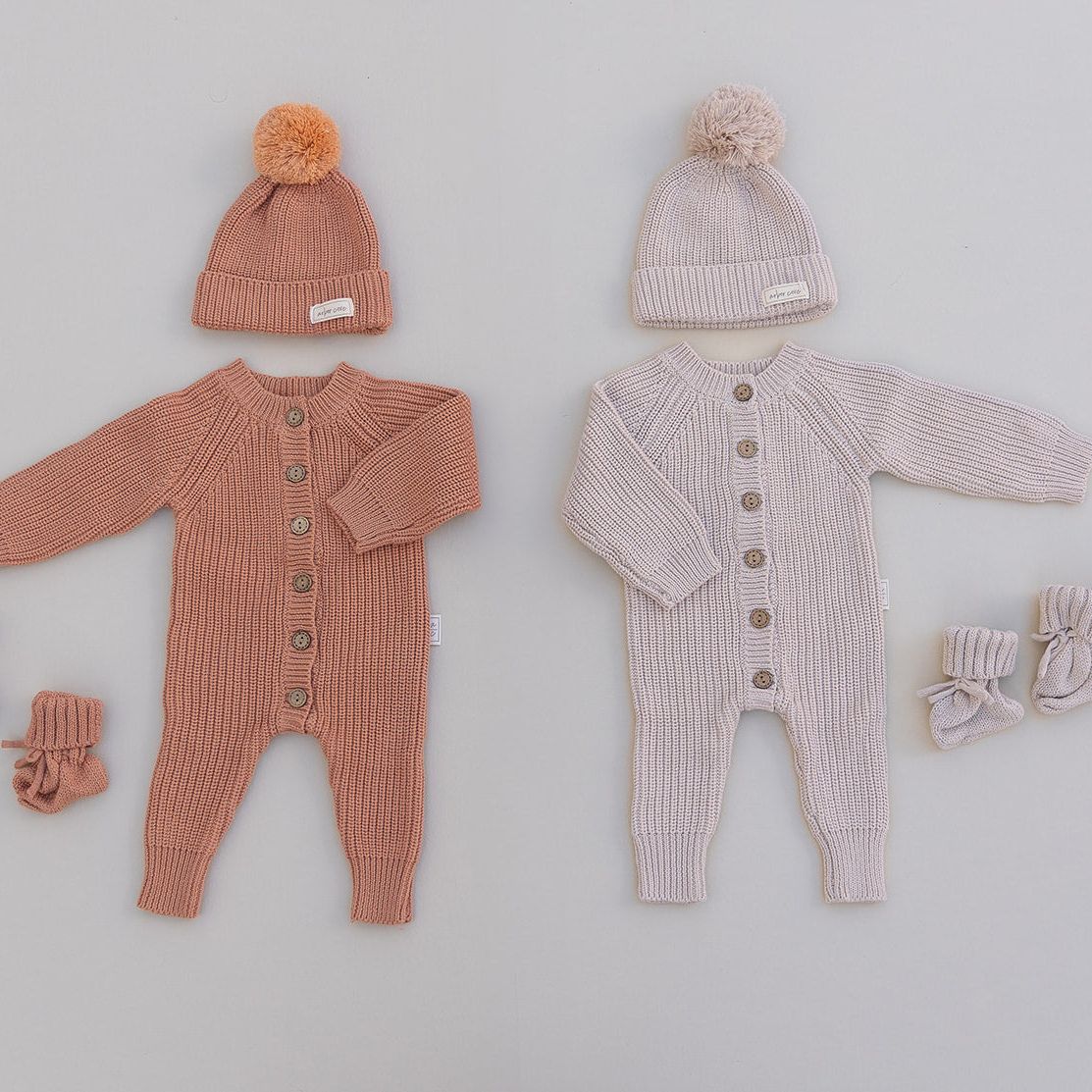 Baby rompers in rust and oat and pom pom beanie and booties on a light gray background
