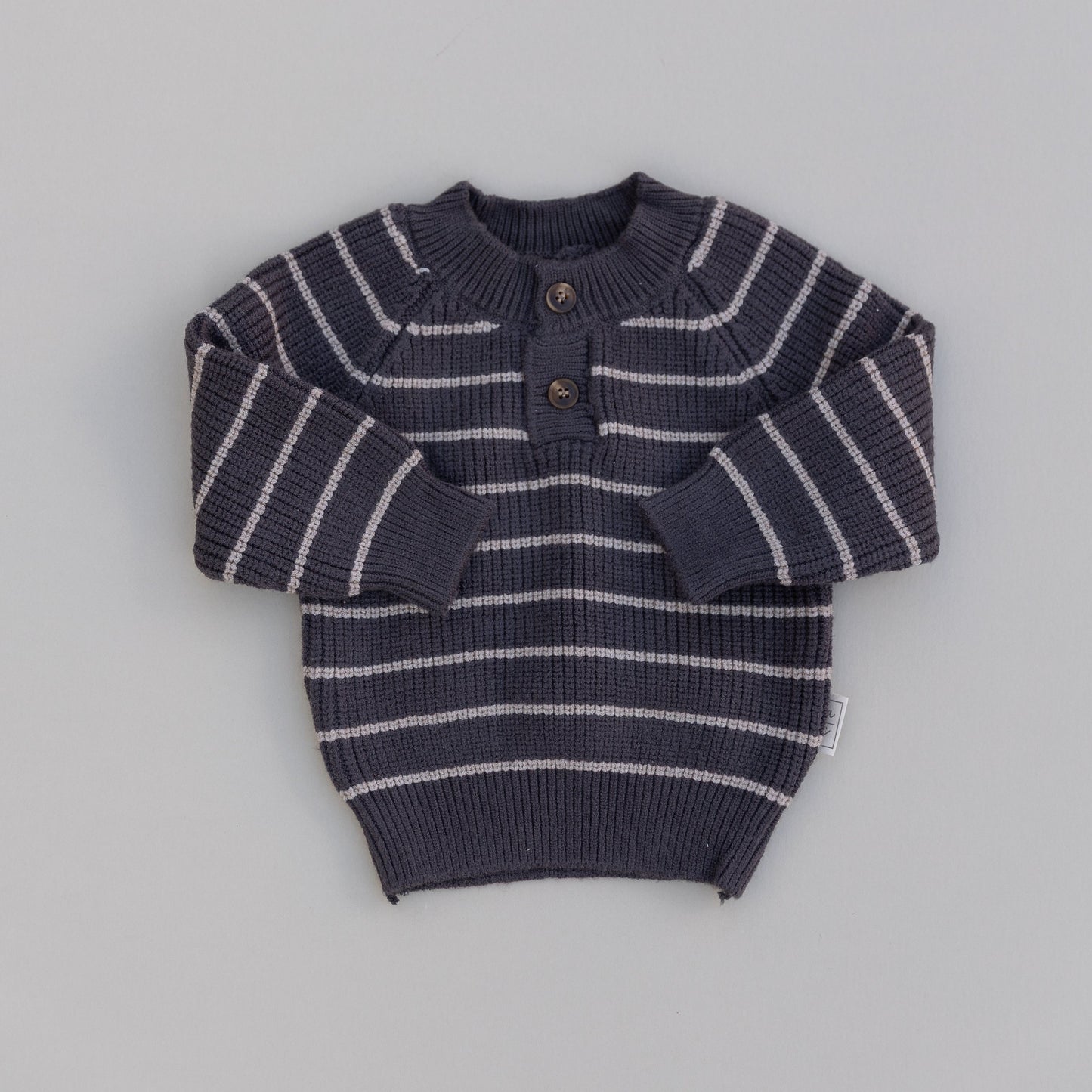 A striped knit button sweater for babies, predominantly in a dark color with light stripes and two front buttons, laid flat on a white background.