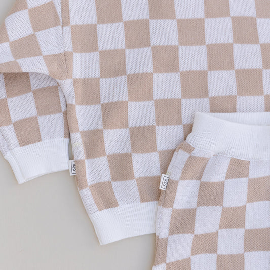 A close-up of a two-piece baby outfit with a checkered long-sleeve sweater and matching bloomers in beige and white colors.