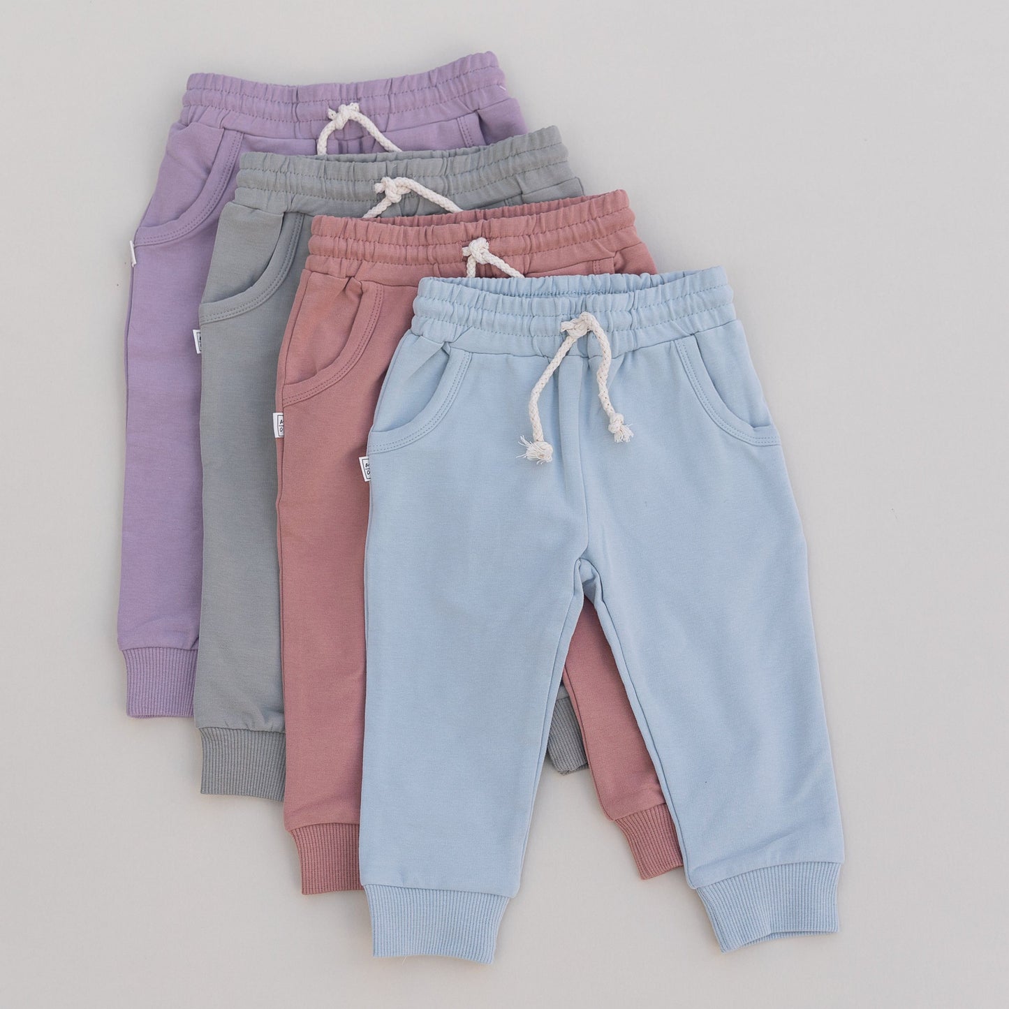 Four pairs of children's jogger pants in purple, green, pink and blue stacked on a light gray background.