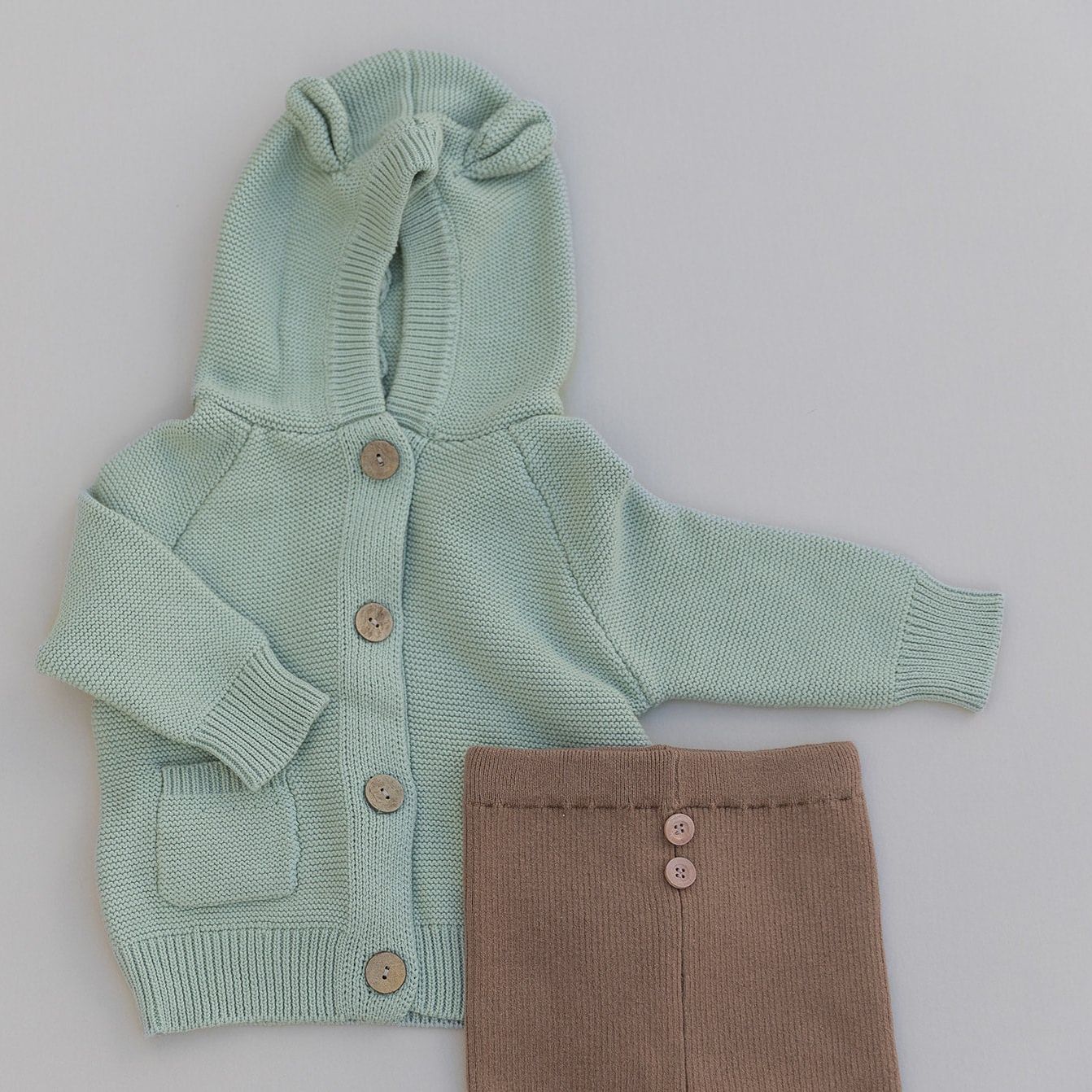 Light green baby cardigan with bear ears and buttons and brown pants with buttons on a light gray background