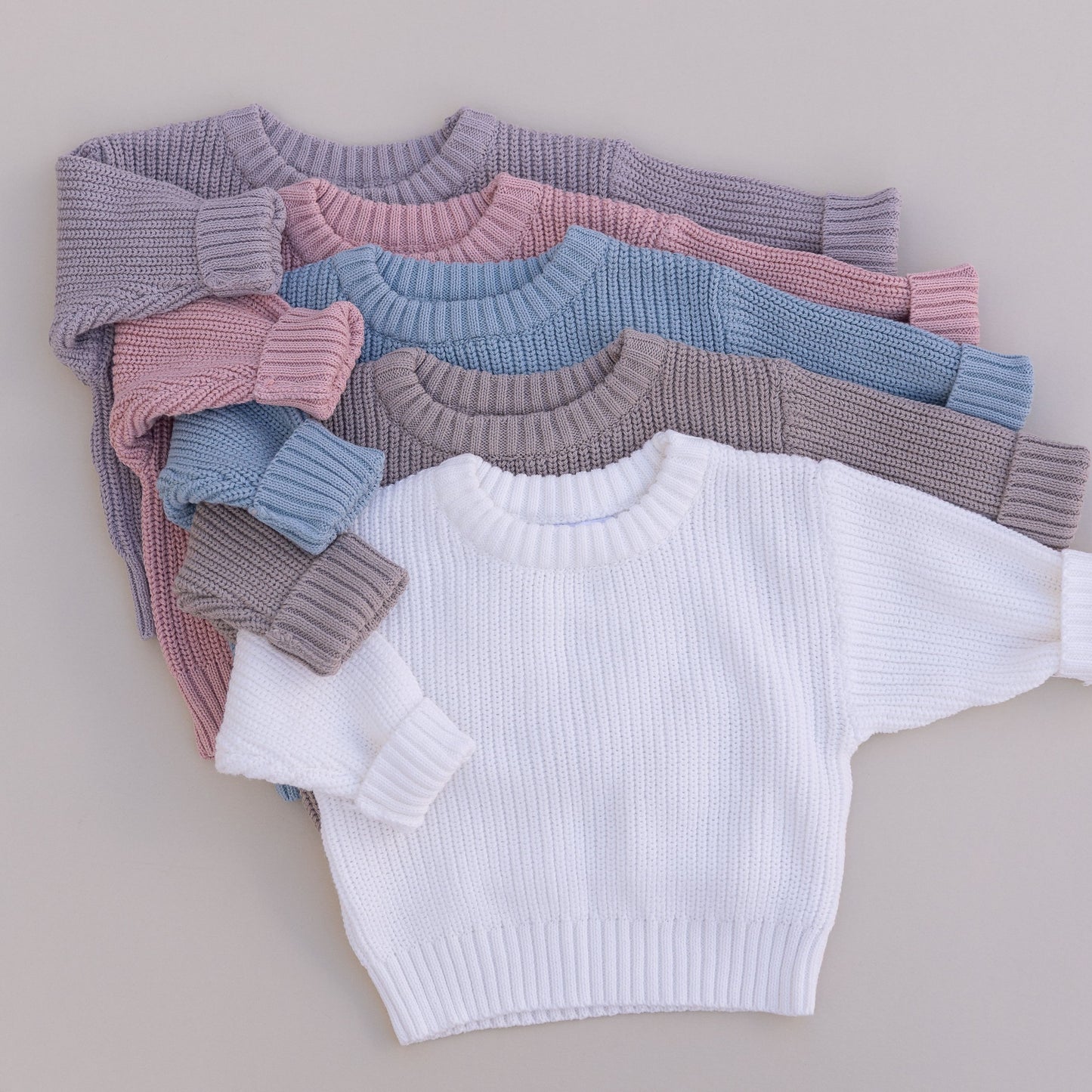 Stack of organic cotton baby sweaters in grey, pink, blue, brown and white on a neutral background