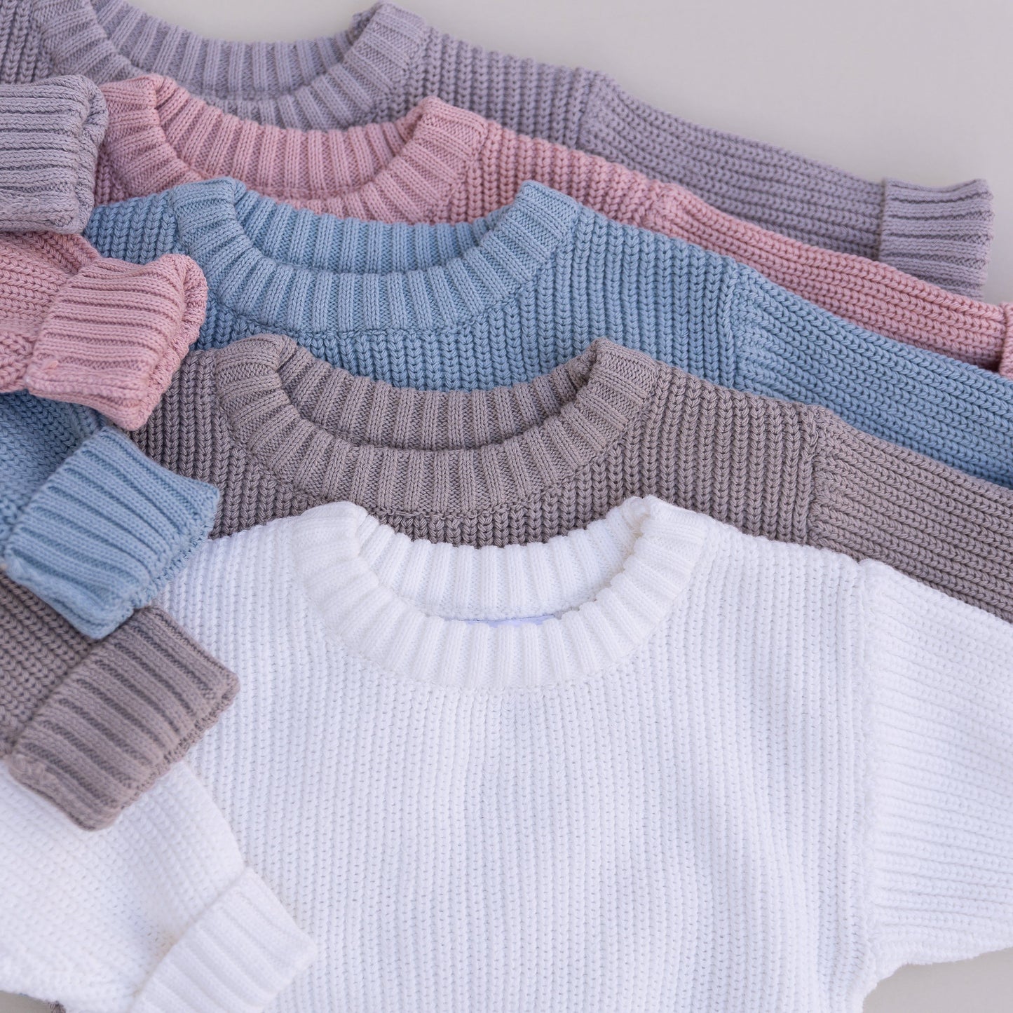 Stack of organic cotton baby sweaters in grey, pink, blue, brown and white on a neutral background