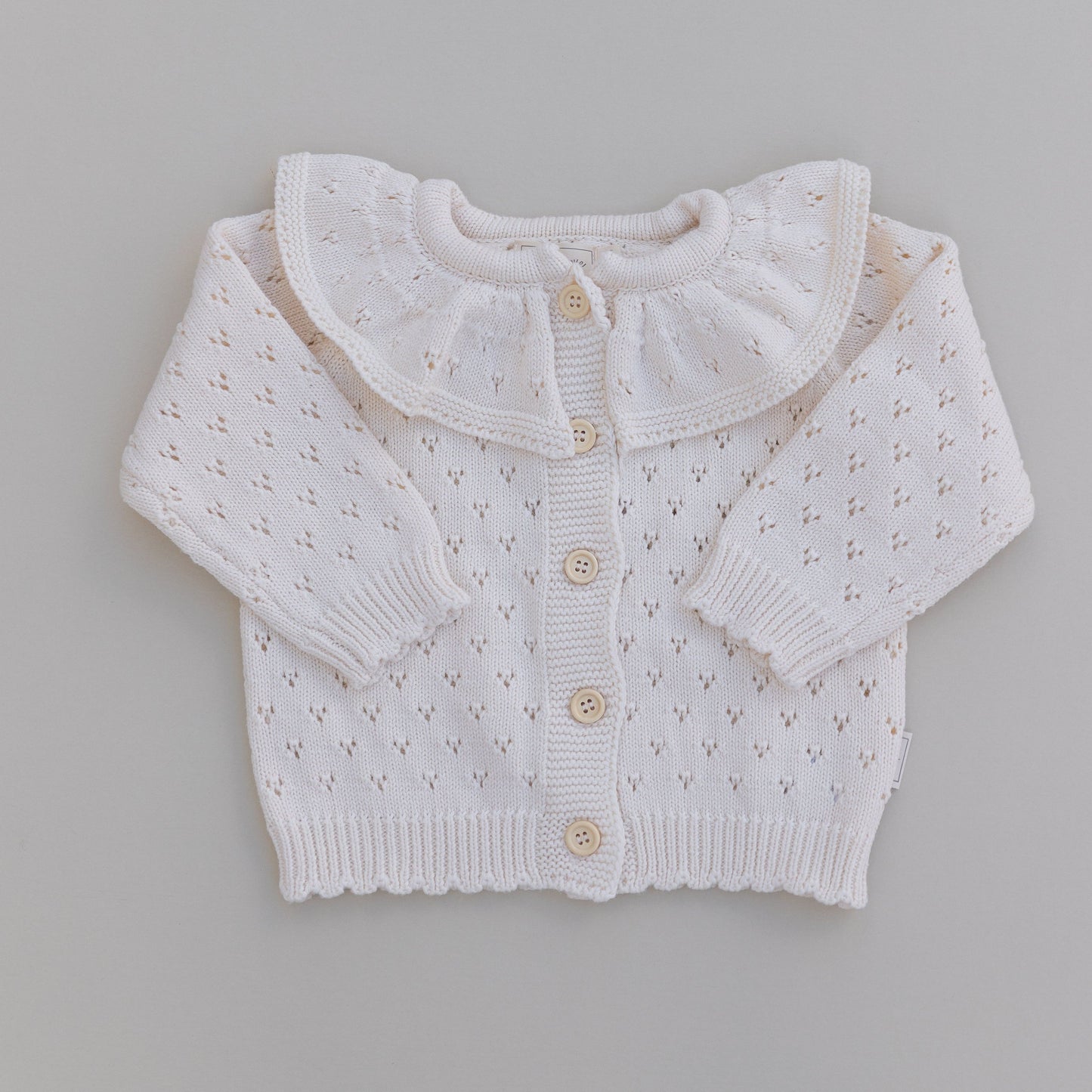 A cream ruffle neck pointelle cardigan with button-down front, laid flat on a neutral background.