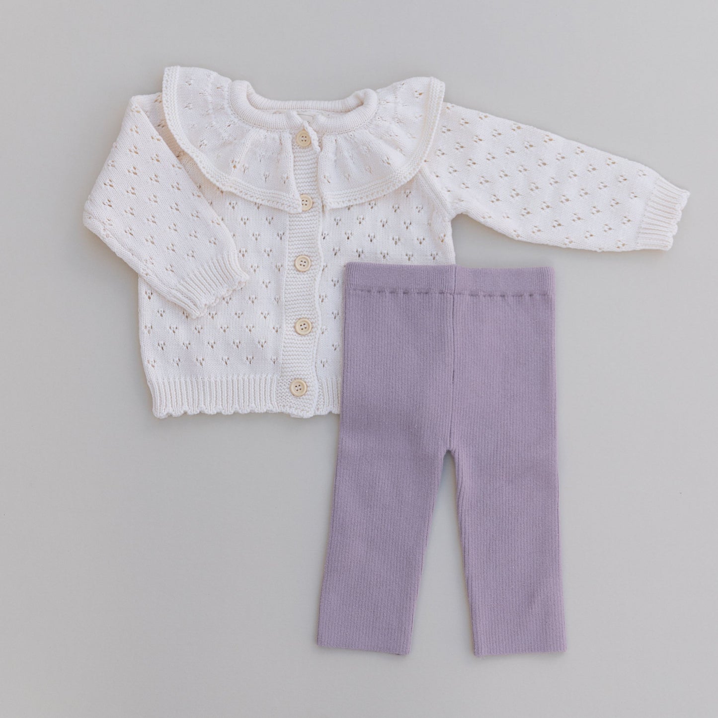 Baby outfit with white ruffled colar pointelle cardigan and purple pants on a gray background