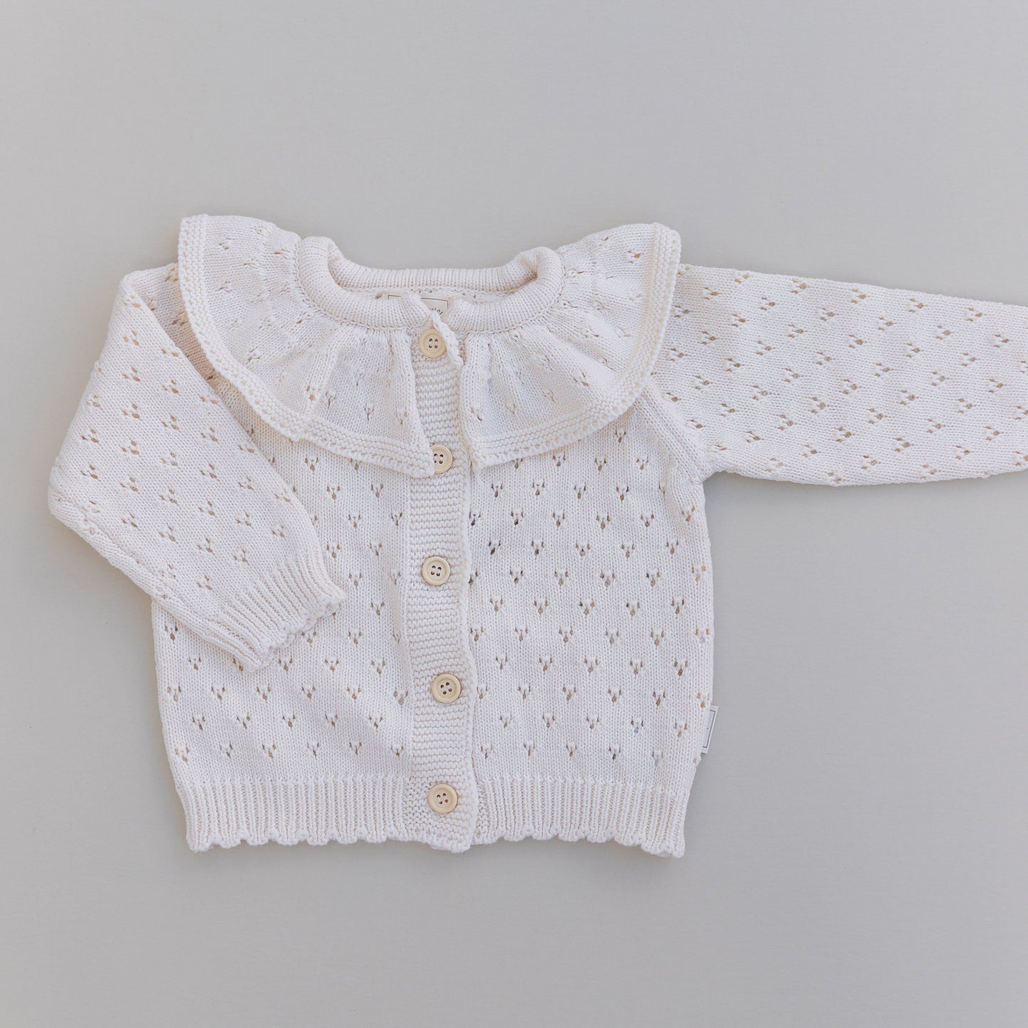 Cream pointelle baby cardigan with a ruffled collar on a gray background
