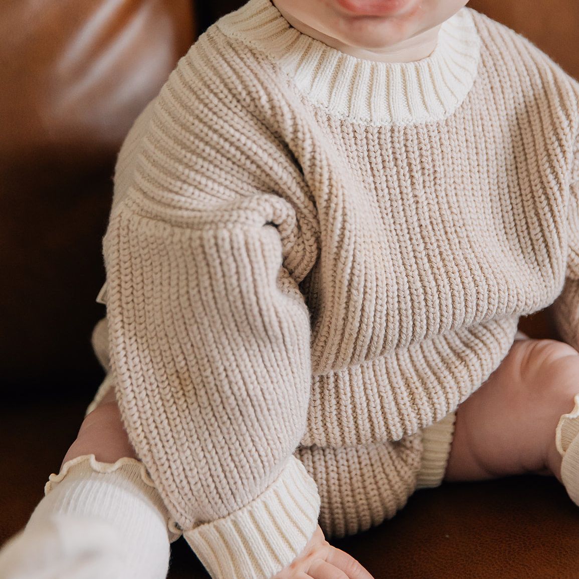 Baby wearing a beige knitted sweater and bloomers sitting on a brown surface.