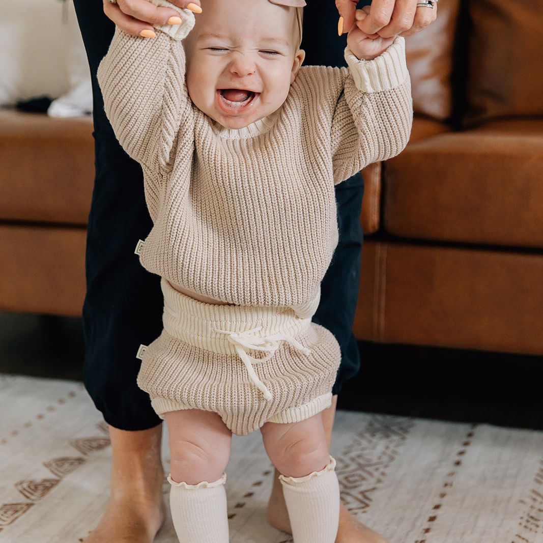 Baby in beige bloomers and sweater outfit with a bow standing on a patterned rug ready for family pictures.