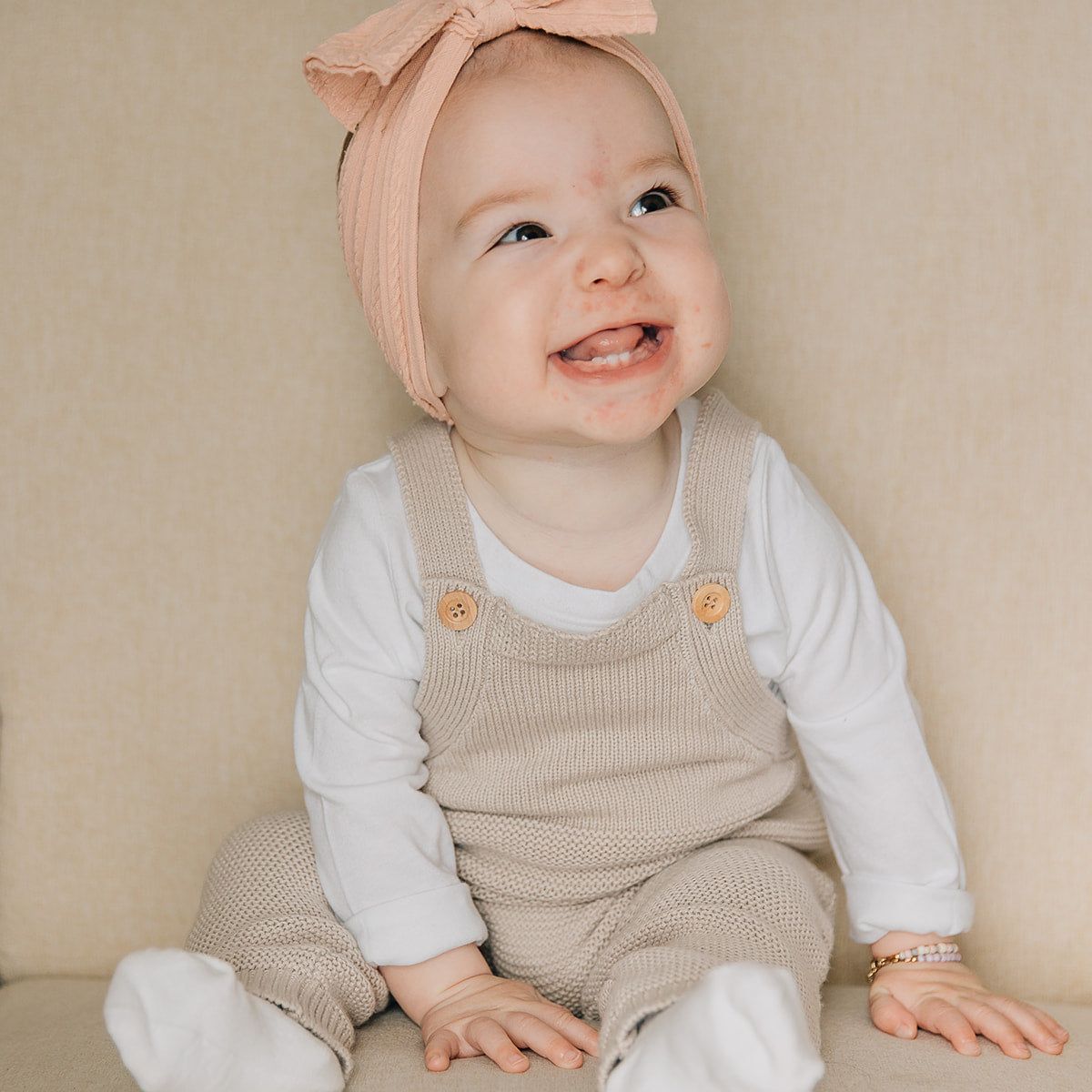 Baby wearing a pink headband and an oat colored knit overalls sitting on a beige couch.