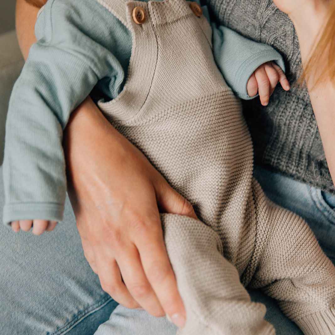 Baby in beige knitted romper being held by an adult, drinking from a bottle.