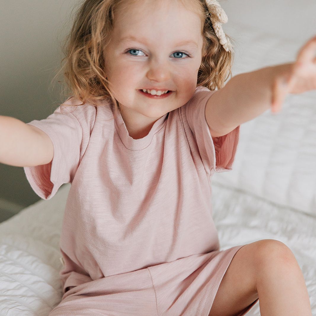Young girl in a pink shorts and shirt sitting on a bed with her arms outstretched.