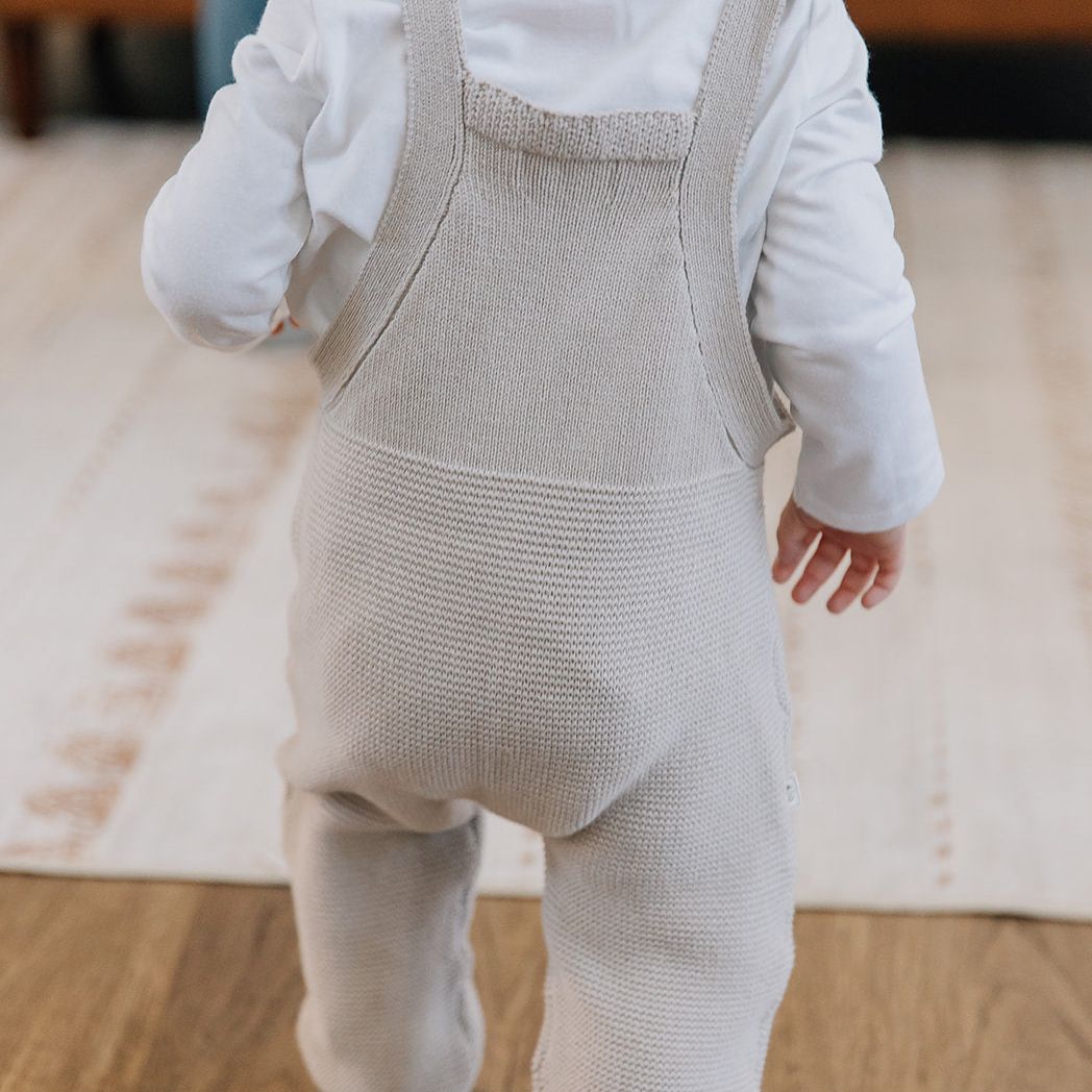 Baby wearing oat colored knit overalls and a white shirt on a wooden floor.