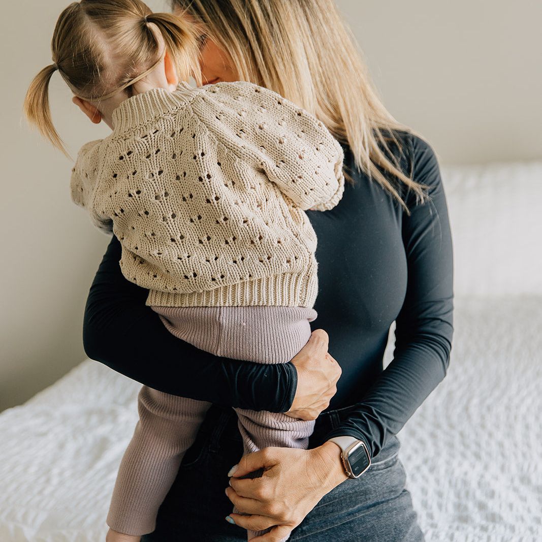 Woman holding a child wearing a beige knitted sweater on a bed.