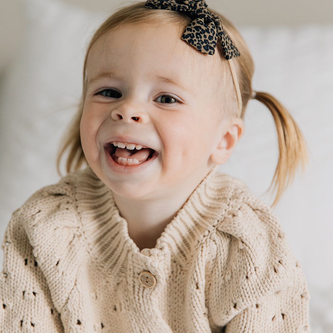 Child wearing a beige knitted cardigan and leopard print headband, sitting on a white surface.