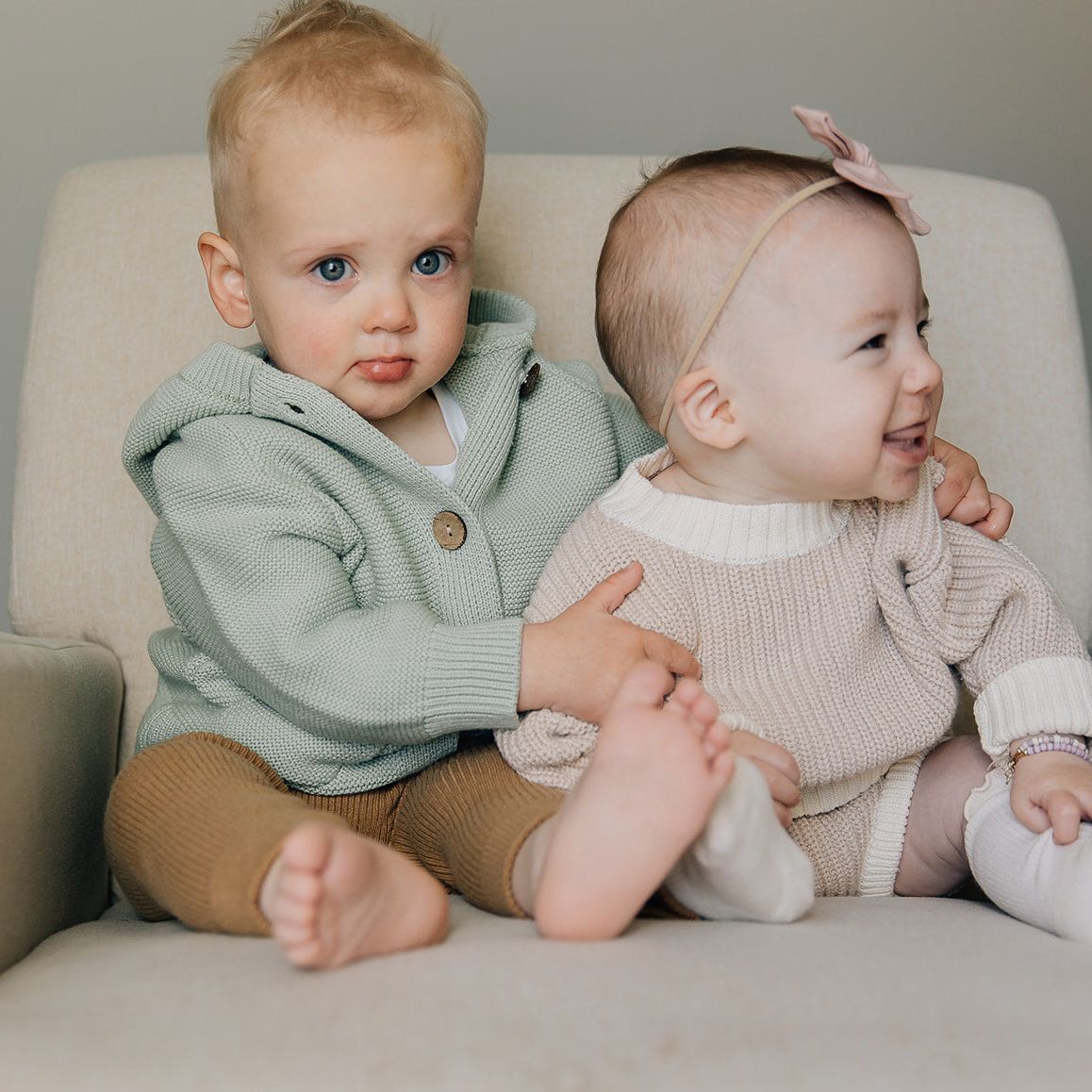 Two young children sitting on a couch, one wearing a green cardigan and the other in a cream sweater and bloomer outfit with a headband.