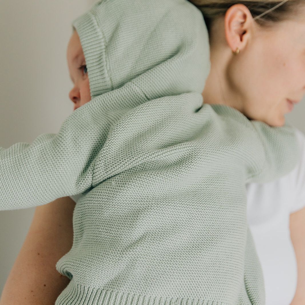 Child wearing a green knitted cardigan with a hood, held by another person.