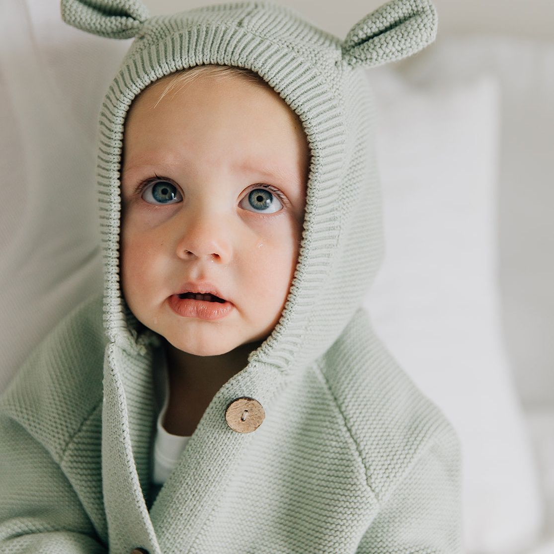 Baby wearing a light green hooded outfit with button details and baby bear ears on a white background