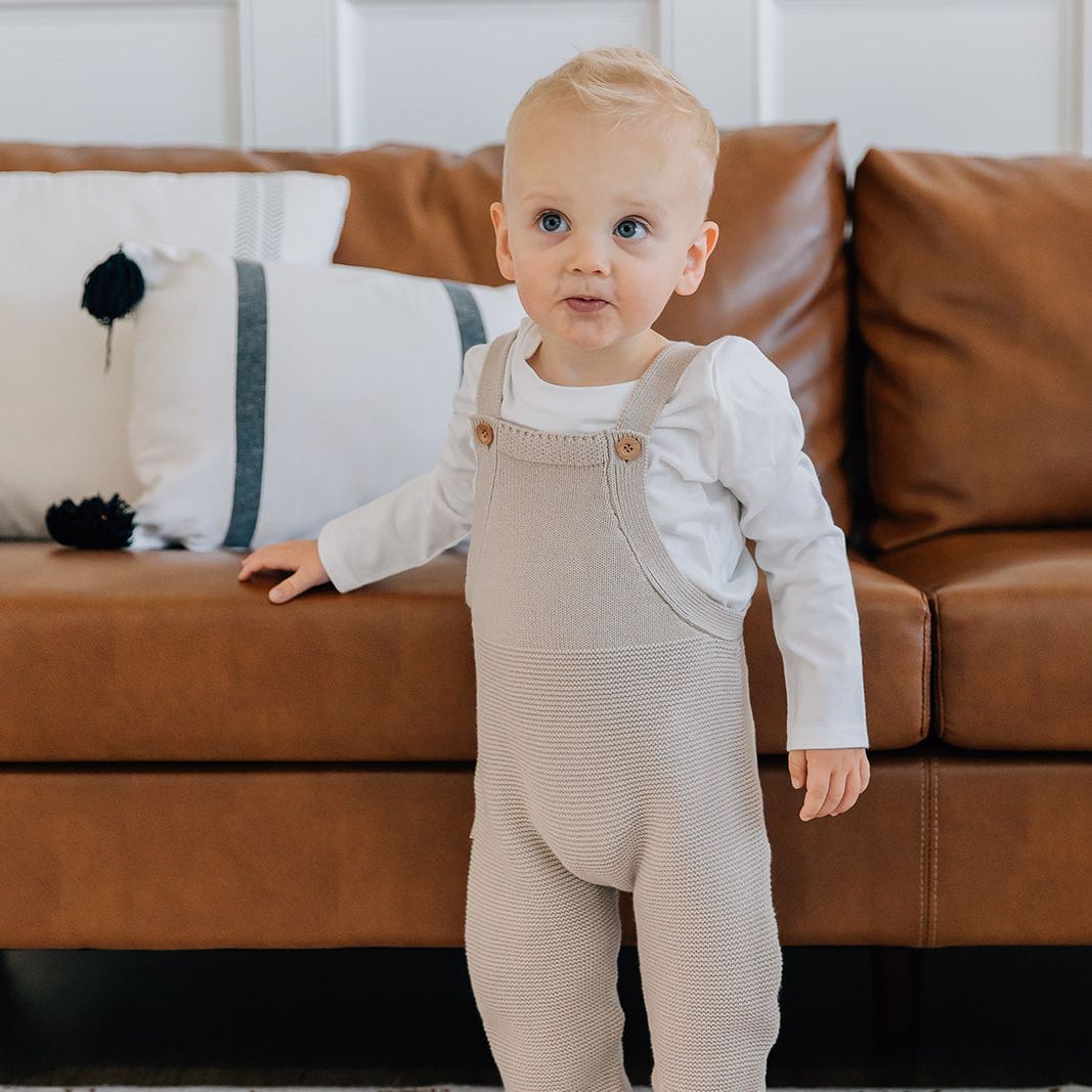 Child wearing oat colored knit overalls standing in a living room with a brown couch and white walls.