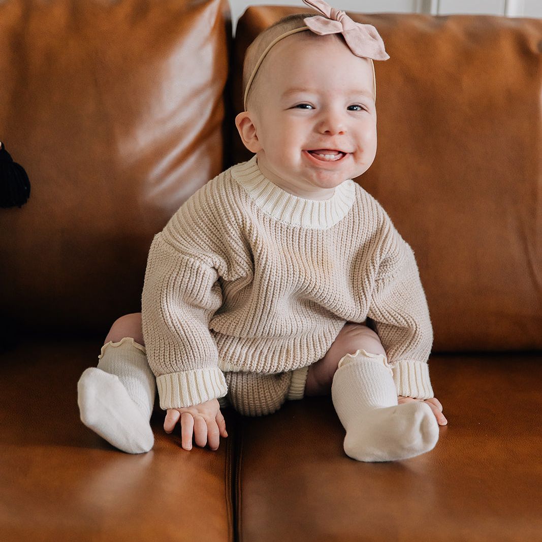 Baby sitting on a brown leather couch wearing a beige sweater and bloomer set and white socks.