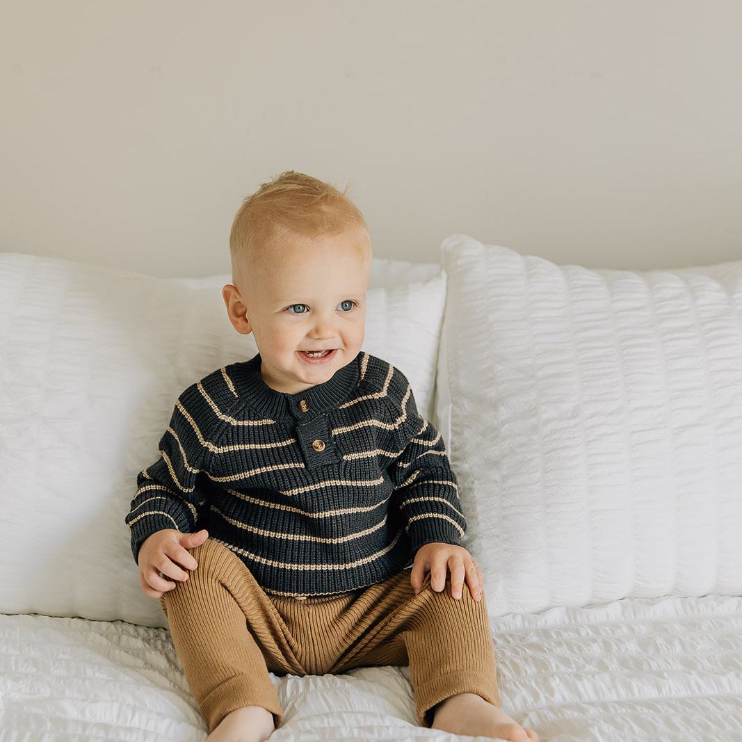 Child wearing a brown sweater with cream stripes and brown pants sitting on a white bed.