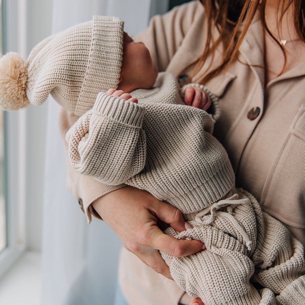 Woman holding a baby wearing a beige knitted sweater and pants set with a pom pom beanie by a window.