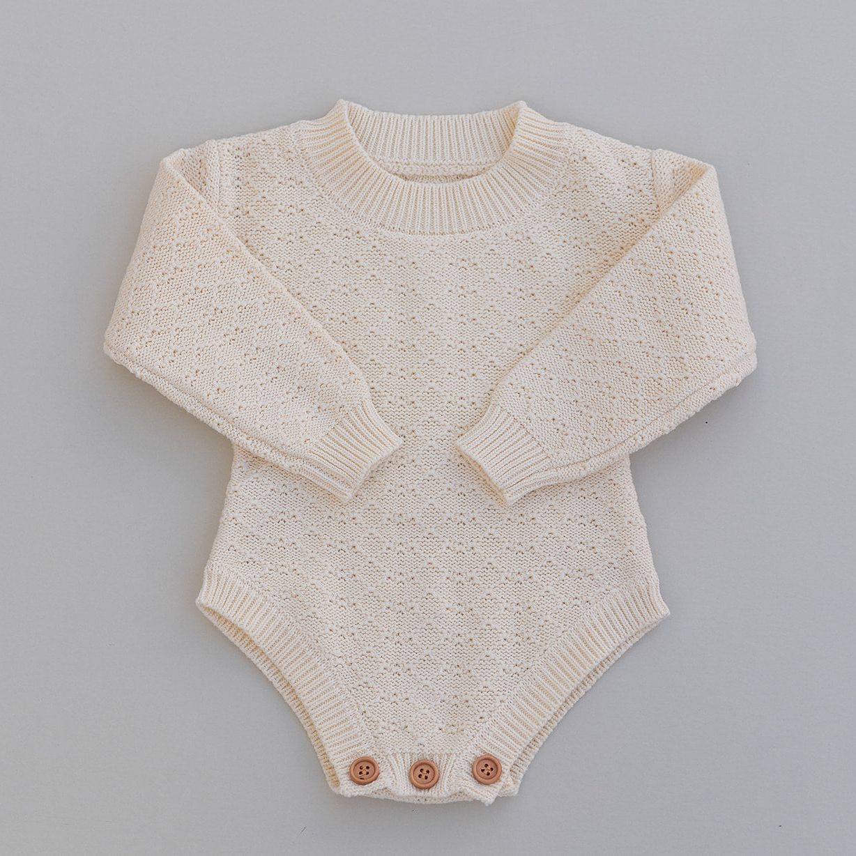 Pointelle knitted long sleeved baby romper in cream with buttons on a gray background