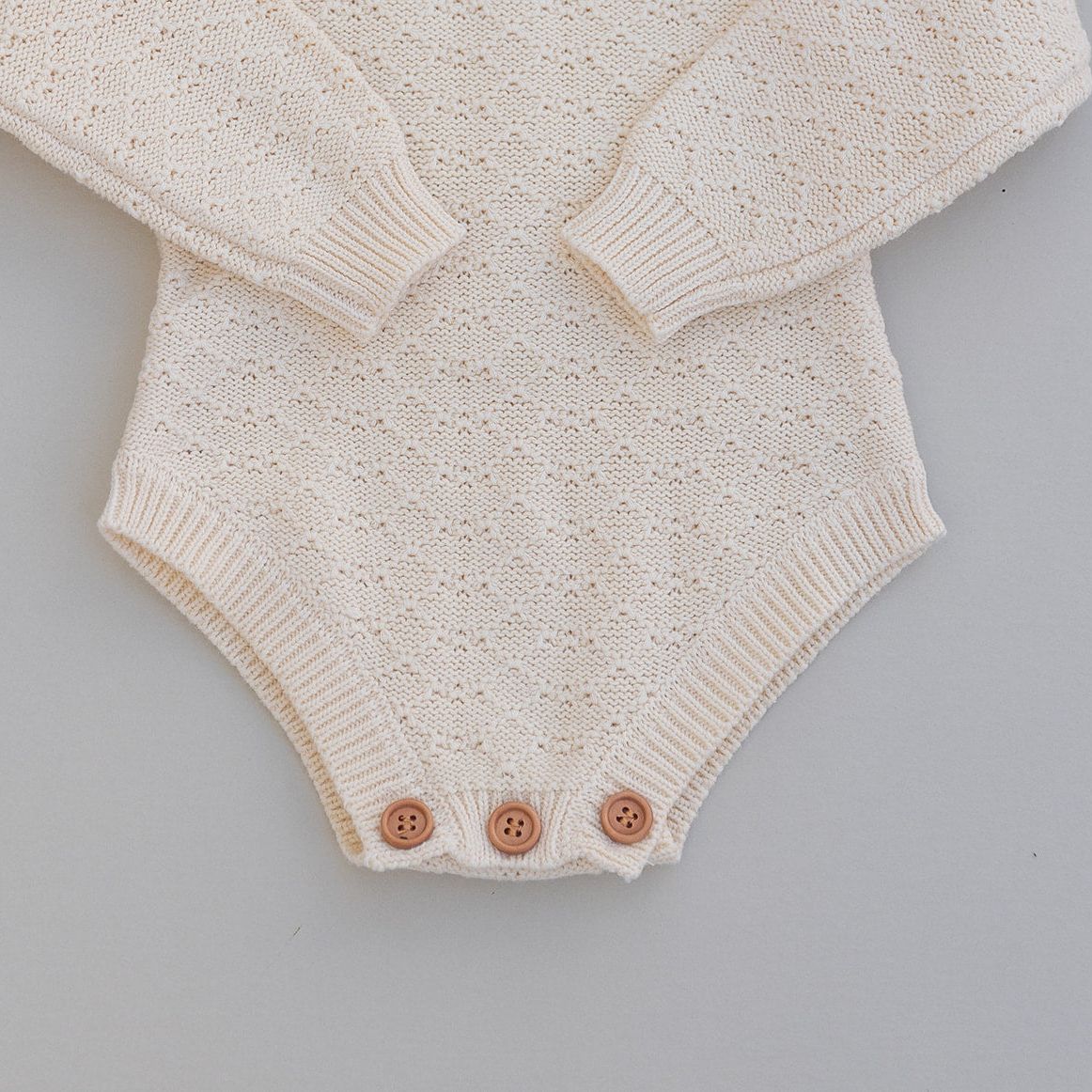Pointelle knitted baby romper in cream with buttons on a gray background