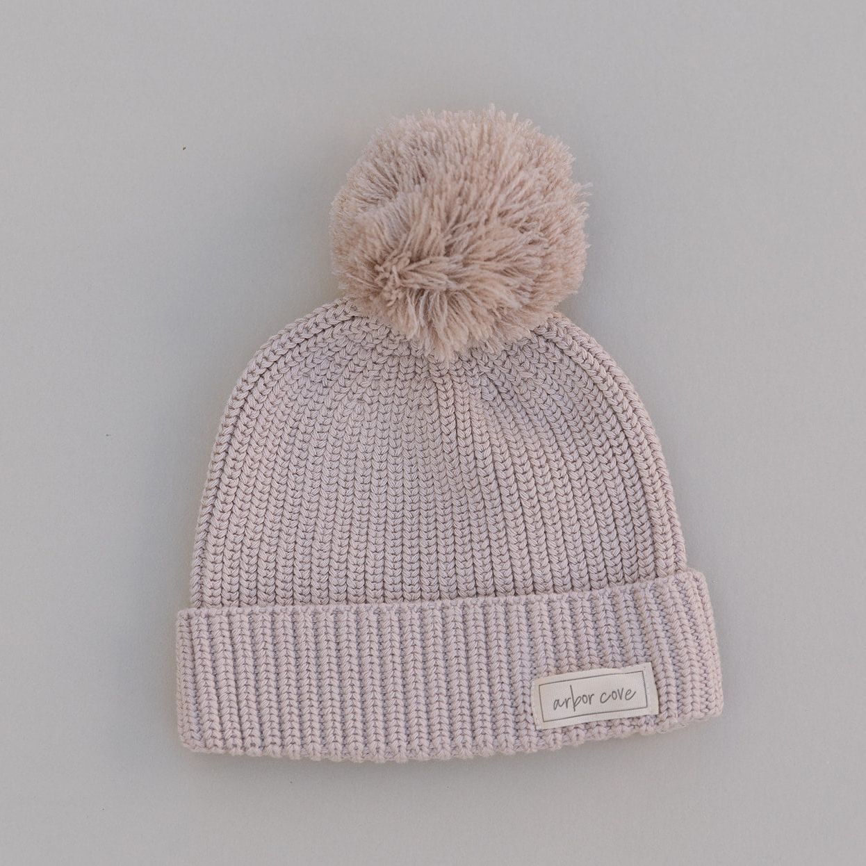 Oat colored knit baby beanie with pom-pom on a light gray background