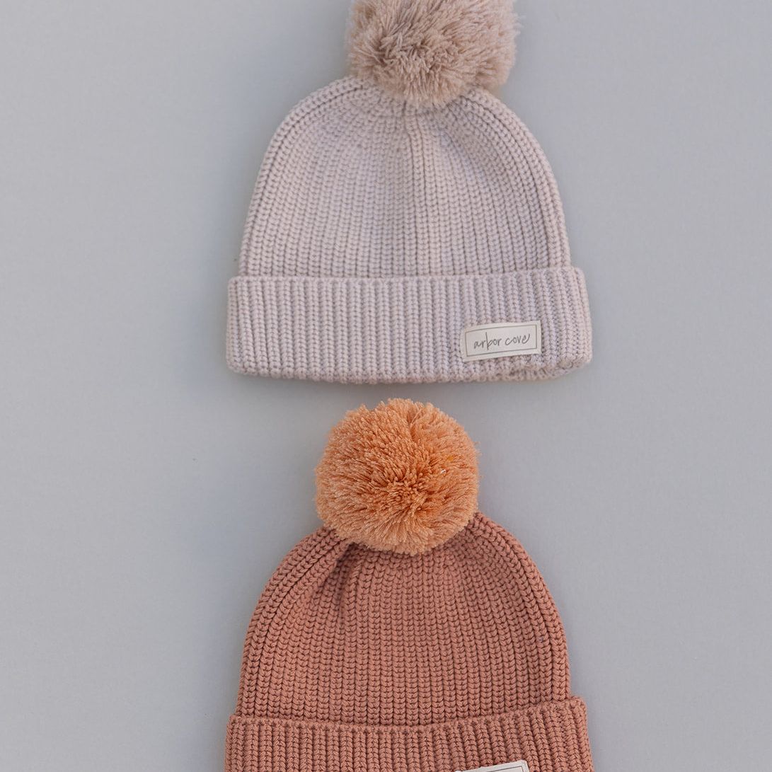 Two knit babyhats with pom-poms, oat and rust colored, on a gray background
