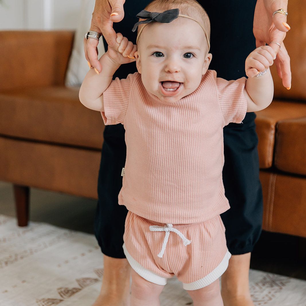 Baby in a pink with white trim ribbed short and t-shirt set being held by an adult in a living room setting