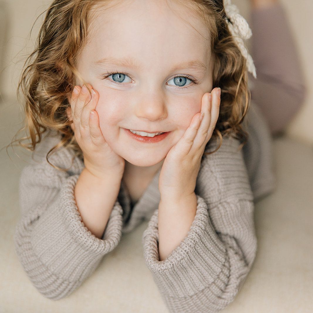 Young girl with curly hair and a flower headband, wearing a gray sweater, sitting on a light-colored surface.