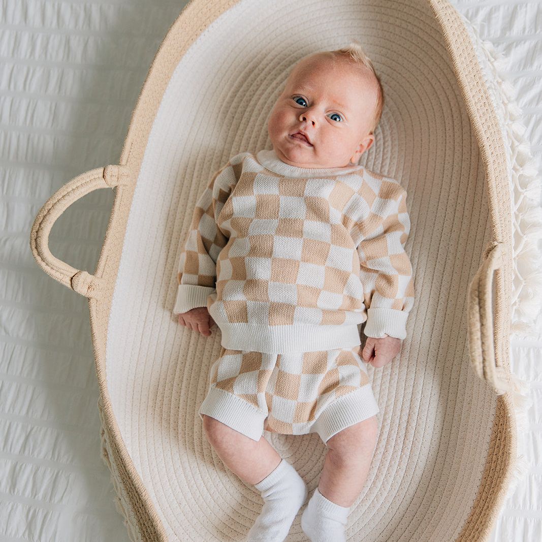 Baby in a two-piece baby outfit with a checkered long-sleeve sweater and matching bloomers in beige and white colors lying in a woven basket on a textured white surface