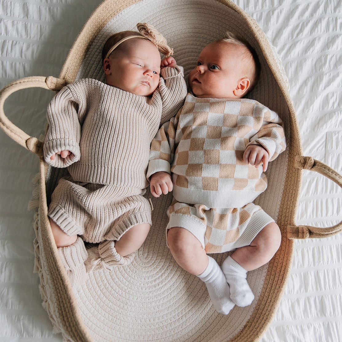 Two babies lying in a woven basket on a textured surface one wearing a cream sweater and bloomer knit set and the other wearing a knit two-piece baby outfit with a checkered long-sleeve sweater and matching bloomers in beige and white colors.