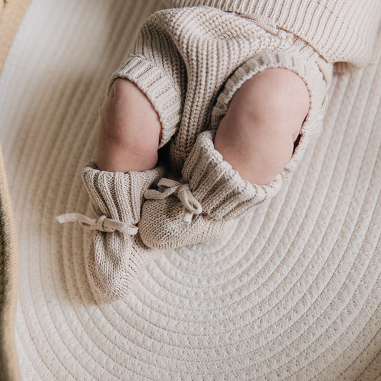 Baby in knitted outfit with bloomers and booties in an oat color lying on a textured surface