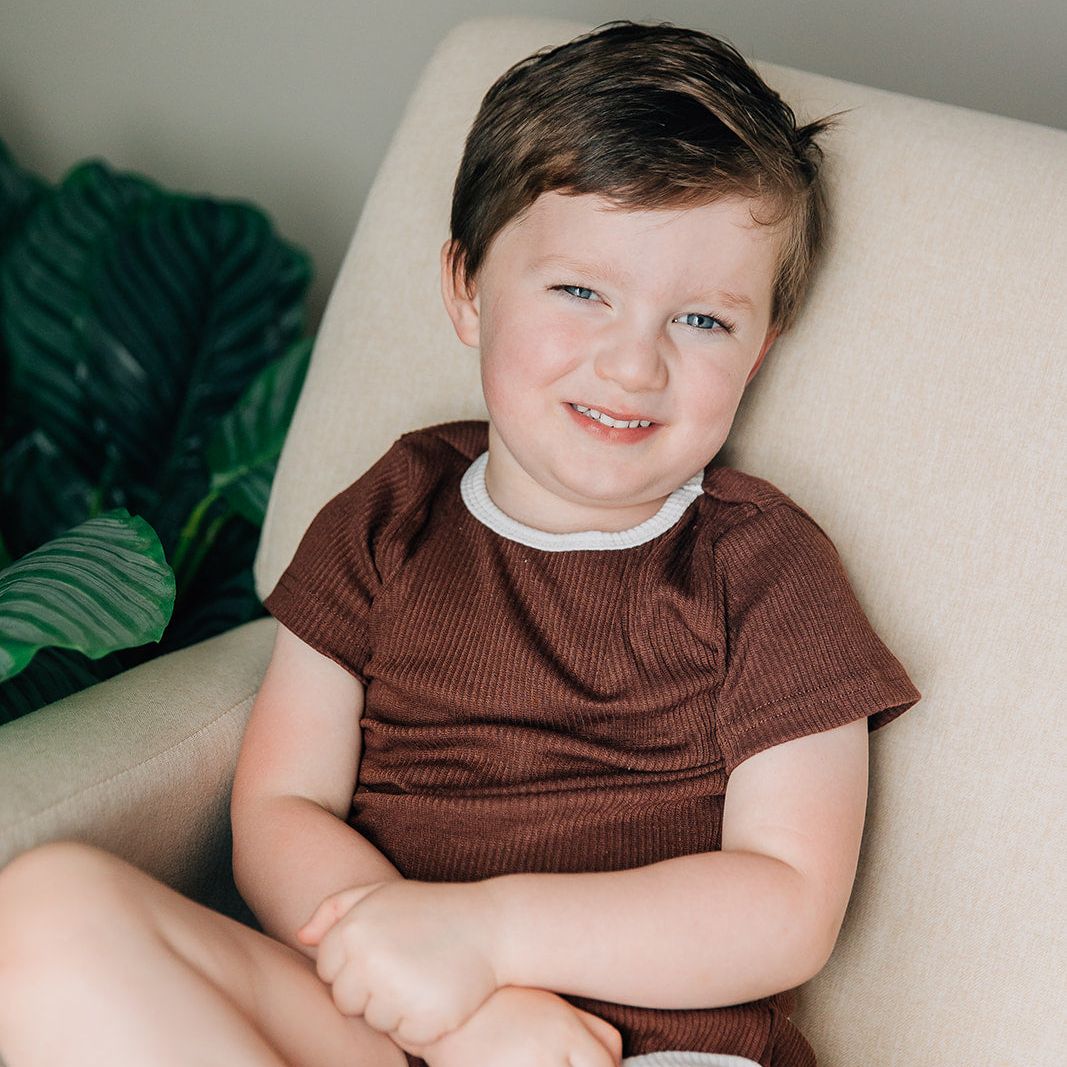 Young boy sitting on a beige chair wearing a brown ribbed shirt with white trim.