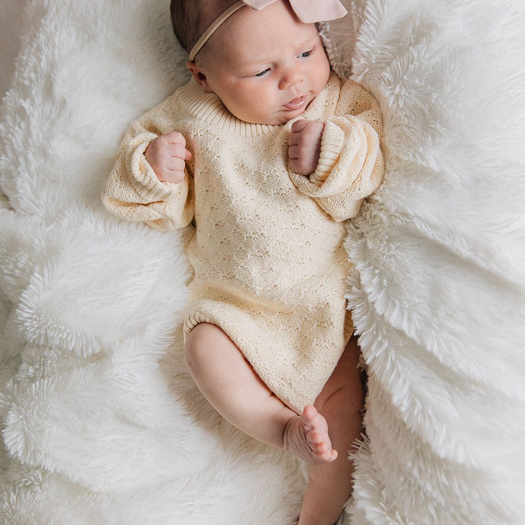 Newborn baby in a cream pointelle knitted baby romper with a pink bow on a white fluffy blanket