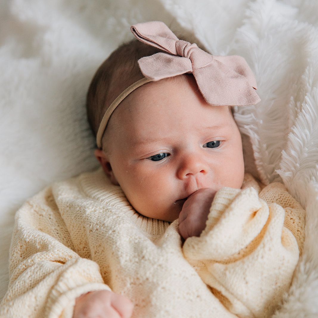 Newborn baby wearing a pointelle knit romper in a cream-colored laying on a blanket with a pink headband.