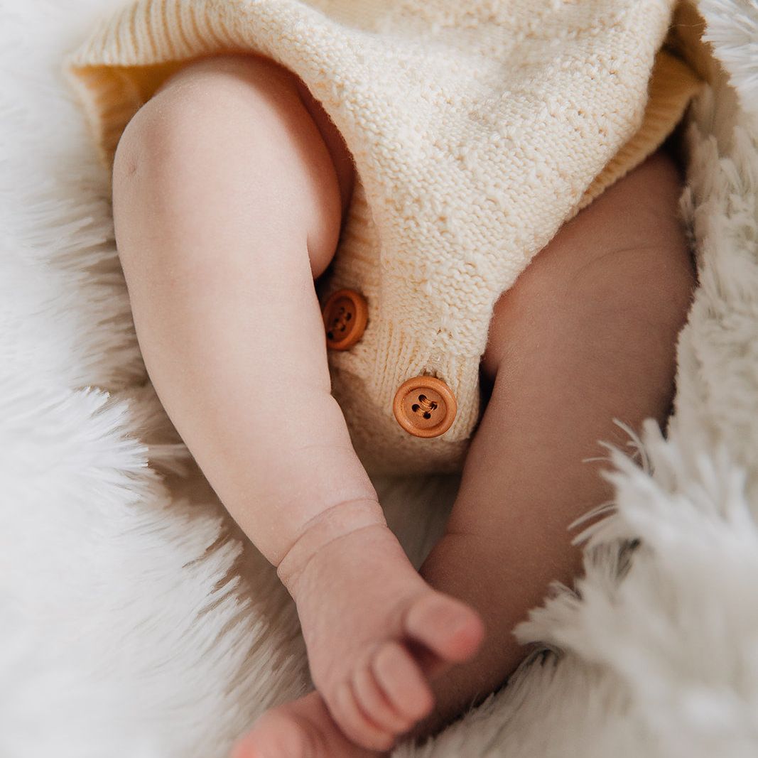 Close-up of a baby's legs wearing a cream colored pointelle knit baby romper buttons on a soft white surface