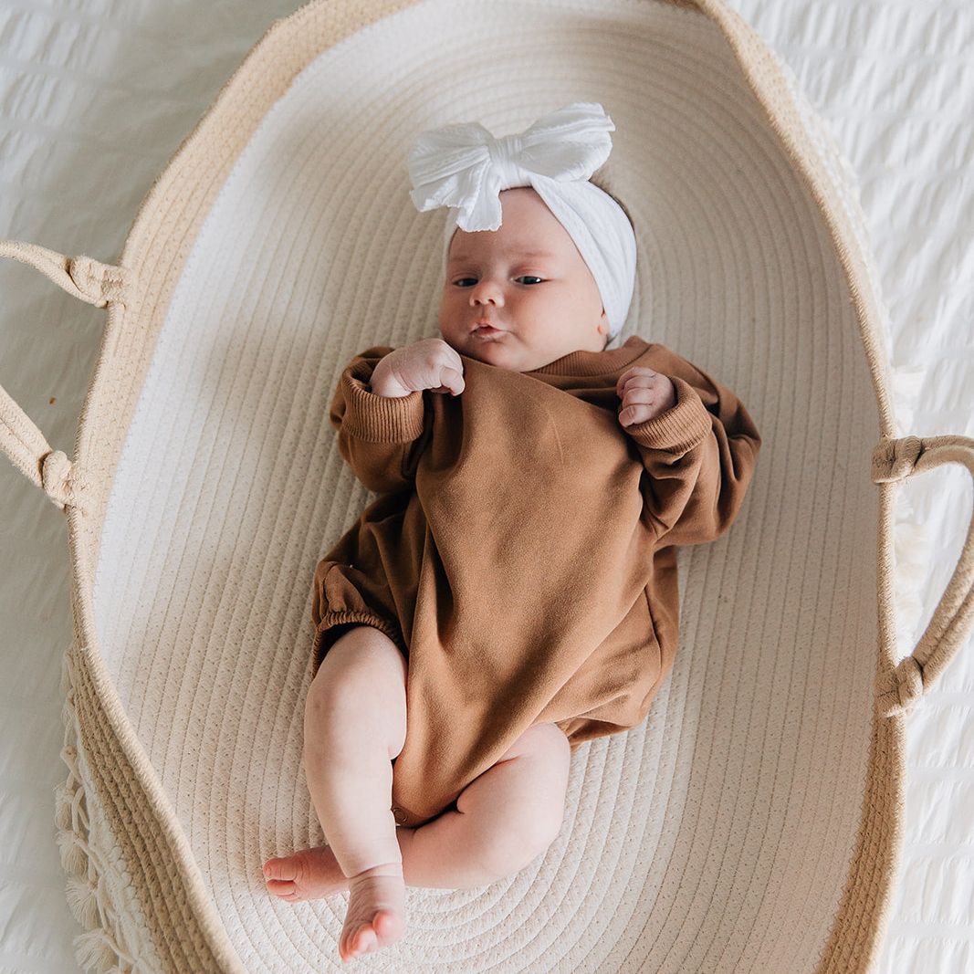 Baby in a brown romper and white headband lying in a woven basket on a textured surface.