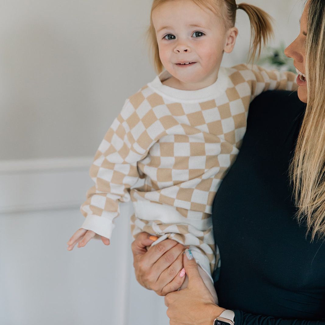 Woman holding a child wearing a two-piece baby outfit with a checkered long-sleeve sweater and matching bloomers in beige and white colors.