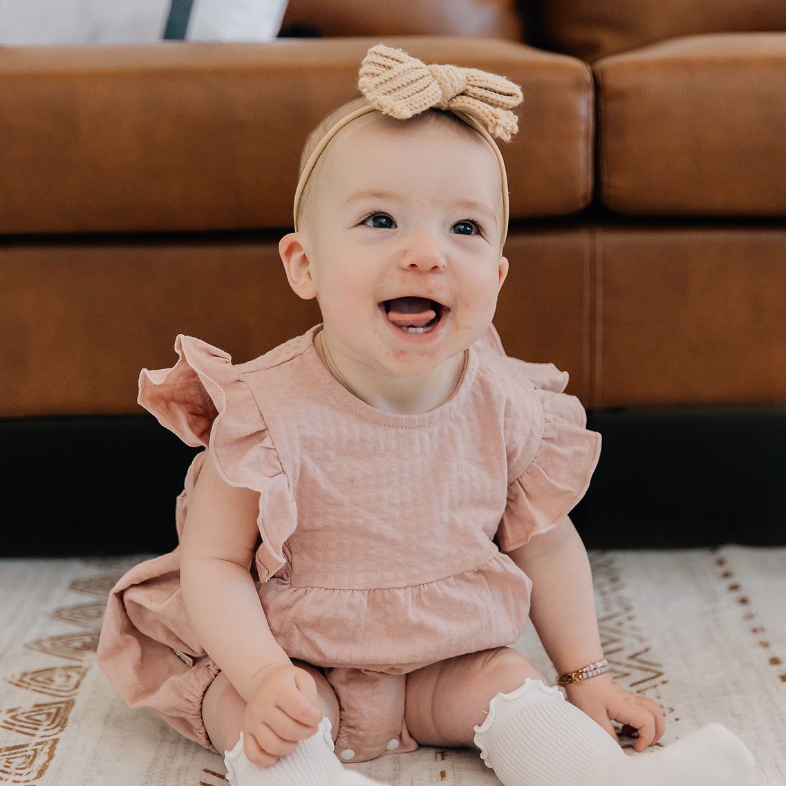 Baby in a pink ruffled bubble romper with ruffled sleeves outfit sitting on a patterned rug.