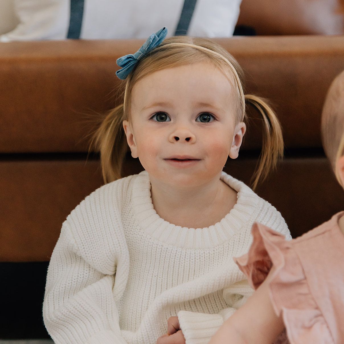 Two young children sitting on a couch, one wearing a white sweater with pigtails and a blue bow, the other partially visible in pink.