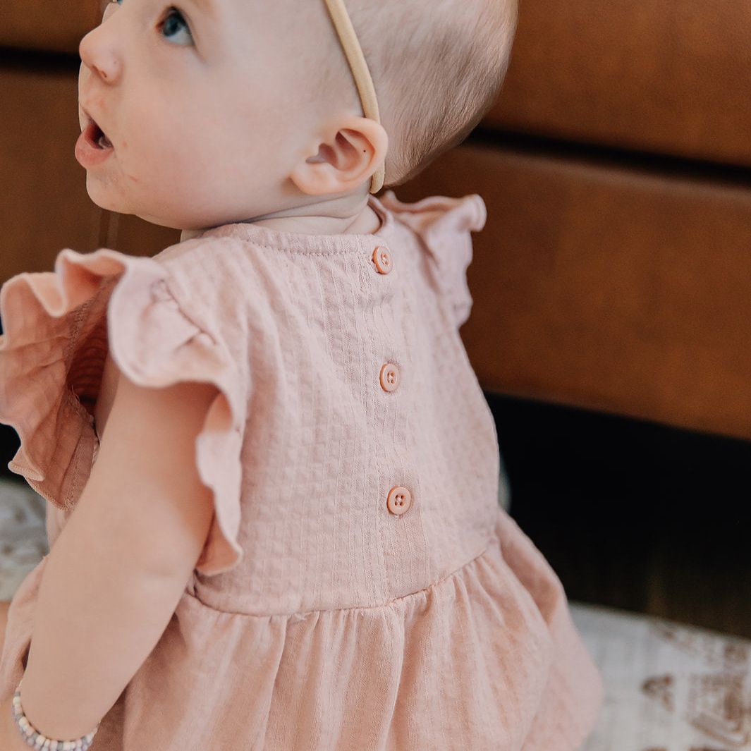 Baby in a pink bubble romper sitting on a patterned surface with a blurred background