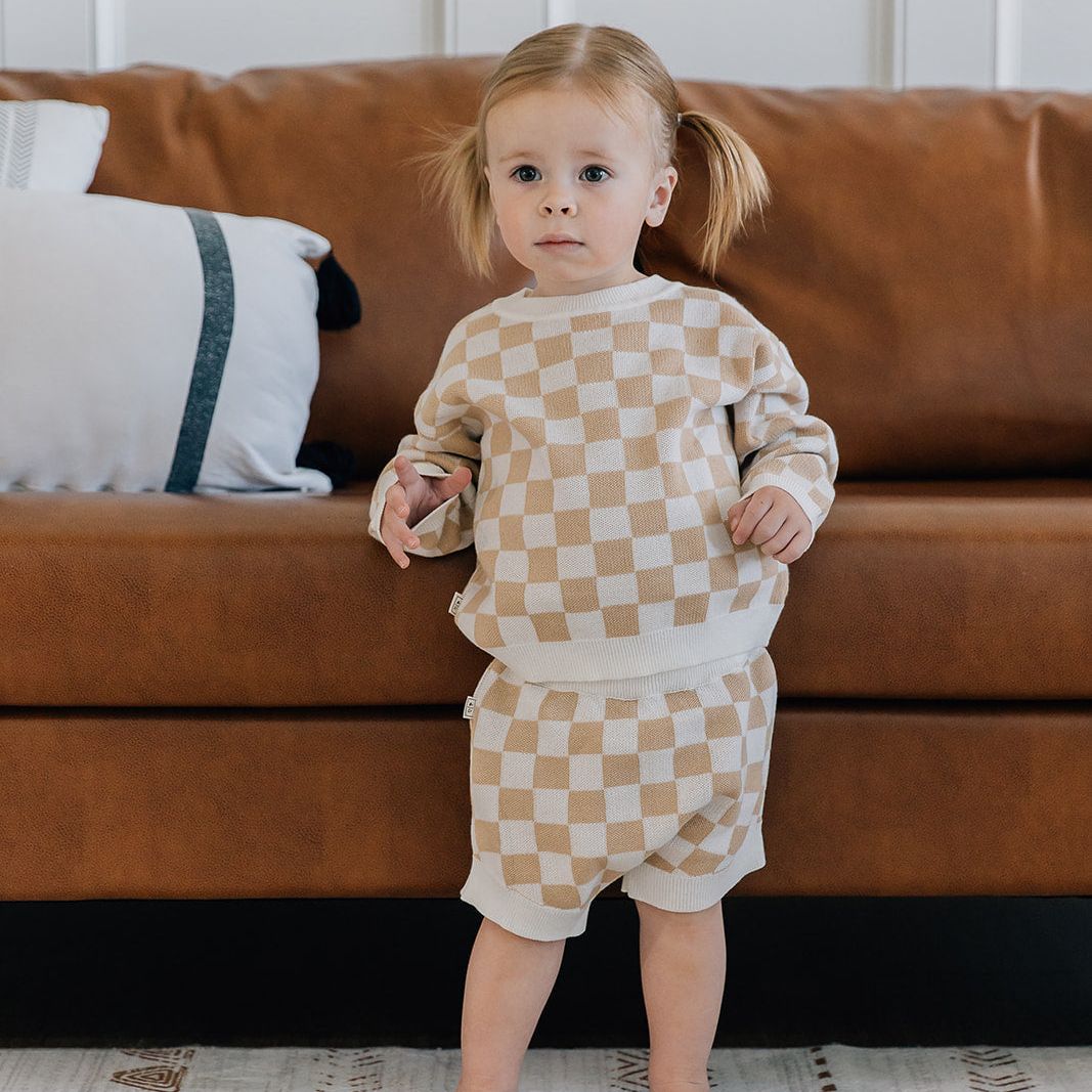 Child wearing a two-piece baby outfit with a checkered long-sleeve sweater and matching bloomers in beige and white colors standing in front of a brown couch.