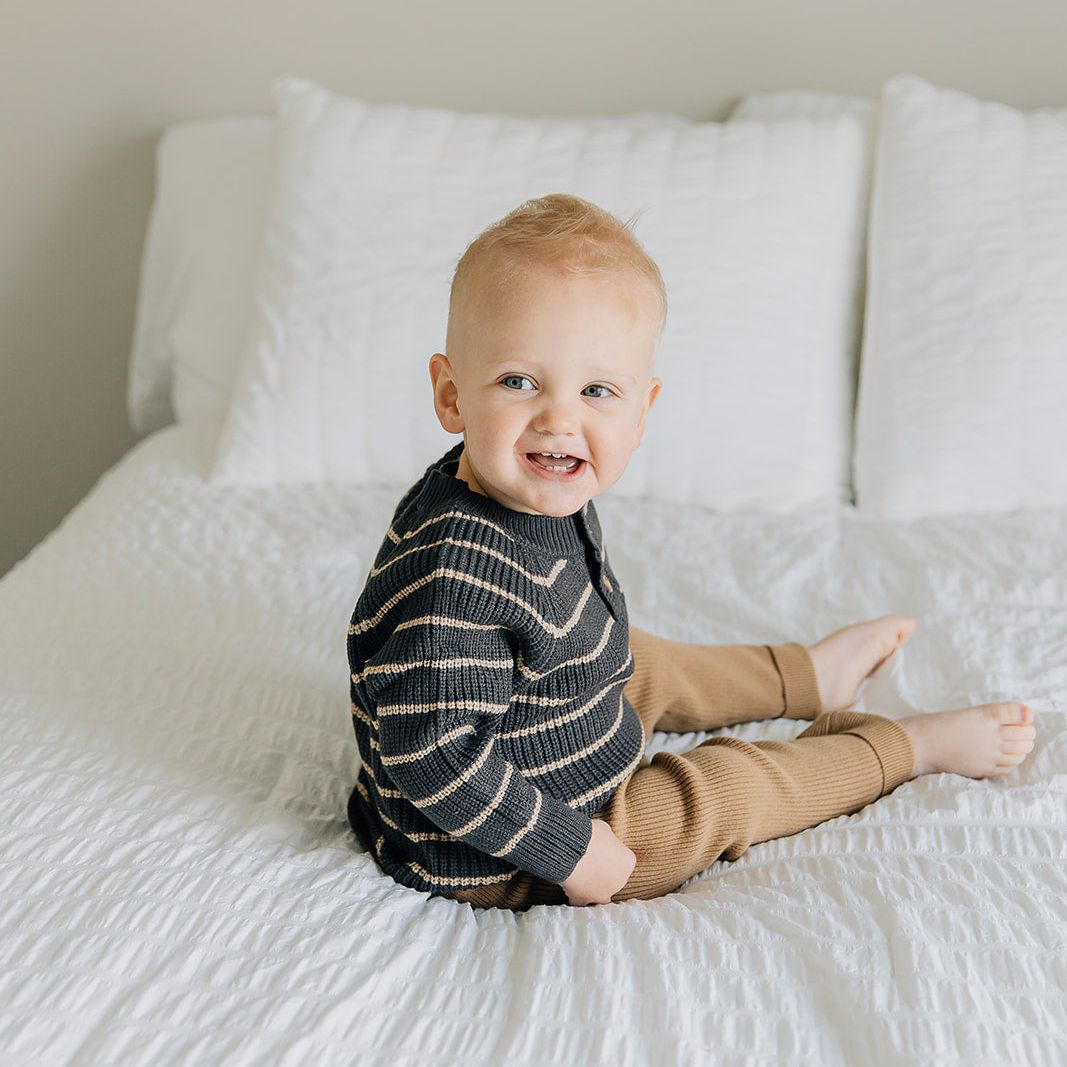 Child sitting on a bed wearing a brown striped knit sweater with cream stripes and brown pants.