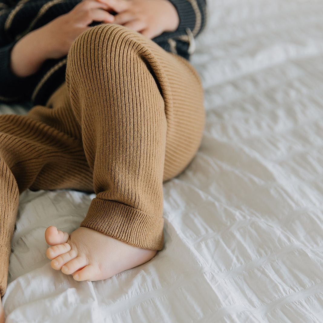 Baby wearing brown ribbed leggings sitting on a white bed.