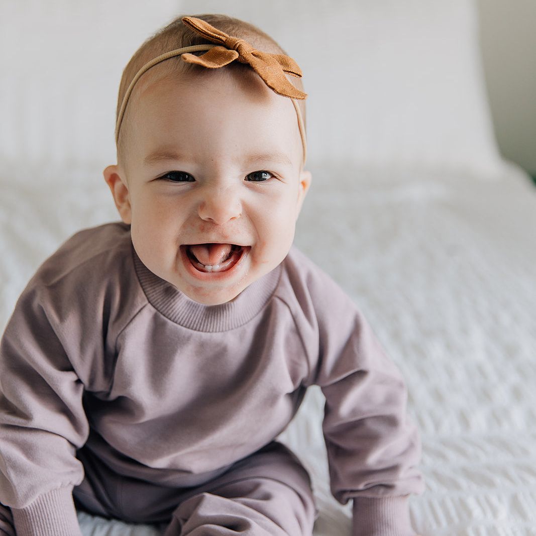 Baby wearing a purple jogger outfit and brown headband sitting on a white bed.