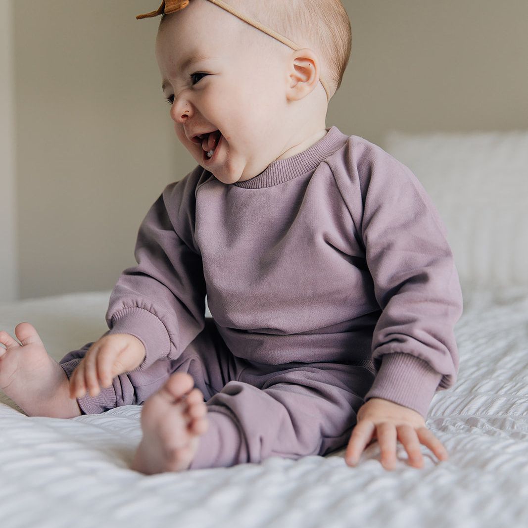 Baby in a purple jogger outfit sitting on a bed with a headband.